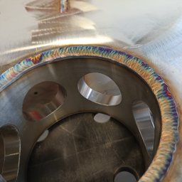 Tigstalmag - Metalowa konstrukcja z otworami, spawana z widocznymi tęczowymi przebarwieniami na spoinie. Detal z bliska, ukazujący precyzję wykonania.