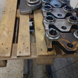 Tigstalmag - Metalowe elementy po obróbce CNC, ułożone na drewnianej palecie. Widoczne ślady obróbki i różnice w wykończeniu powierzchni. Elementy o różnych kształtach i rozmiarach.