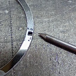 Tigstalmag - Metalowy pierścień z wyciętym otworem leży na metalowej powierzchni obok rysika. Detal obróbki CNC lub spawania, widoczne ślady pracy.