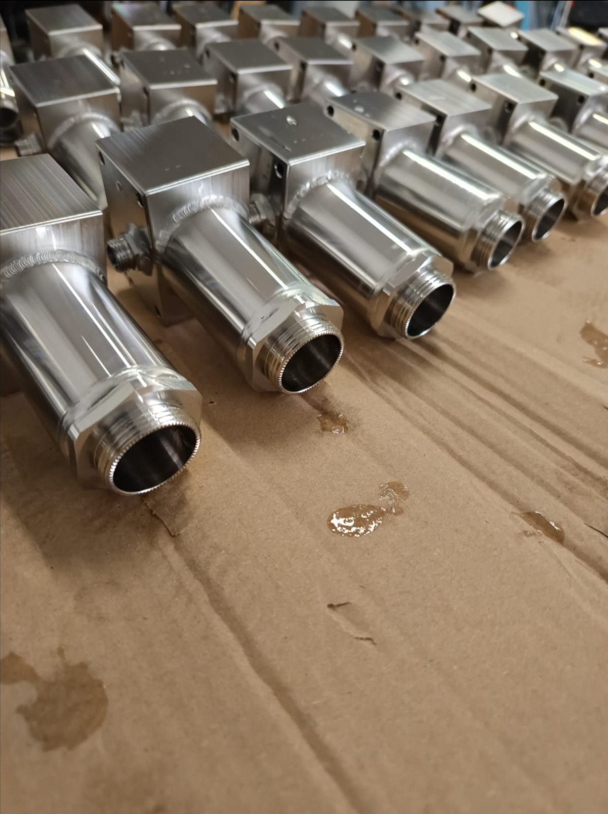 Rząd precyzyjnie wykonanych, metalowych elementów z gwintem, ułożonych na kartonie. Widoczne ślady obróbki i połyskliwa powierzchnia.