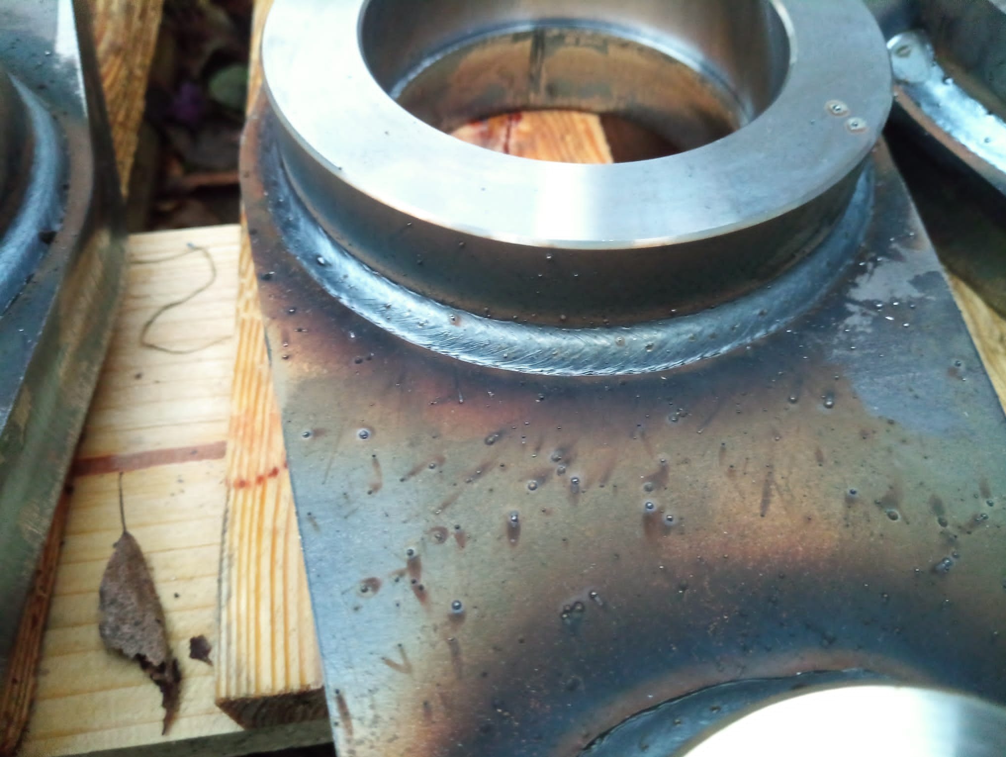 Metalowy element po spawaniu z widocznymi śladami obróbki termicznej i spoinami, leżący na drewnianej powierzchni. Detal z bliska, ukazujący precyzję wykonania.