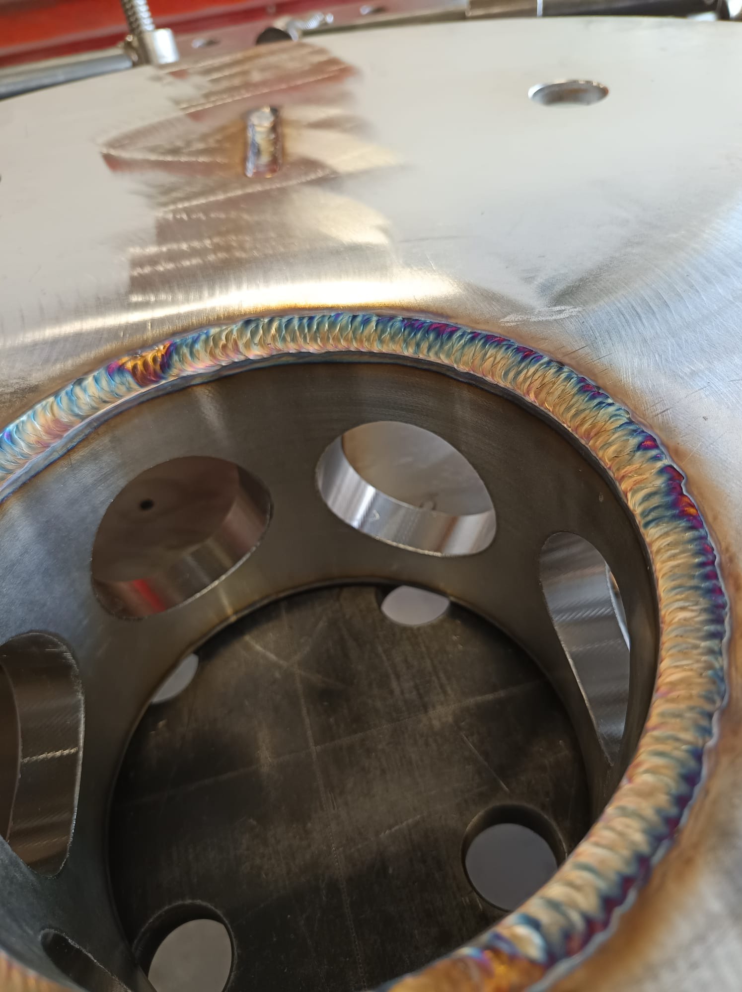 Metalowa konstrukcja z otworami, spawana z widocznymi tęczowymi przebarwieniami na spoinie. Detal z bliska, ukazujący precyzję wykonania.