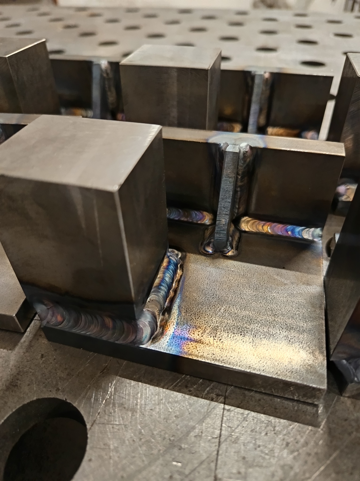 Precyzyjne spawy łączące metalowe elementy w prostopadłościenną konstrukcję. Widoczne tęczowe przebarwienia na spoinach, świadczące o wysokiej temperaturze i jakości wykonania.