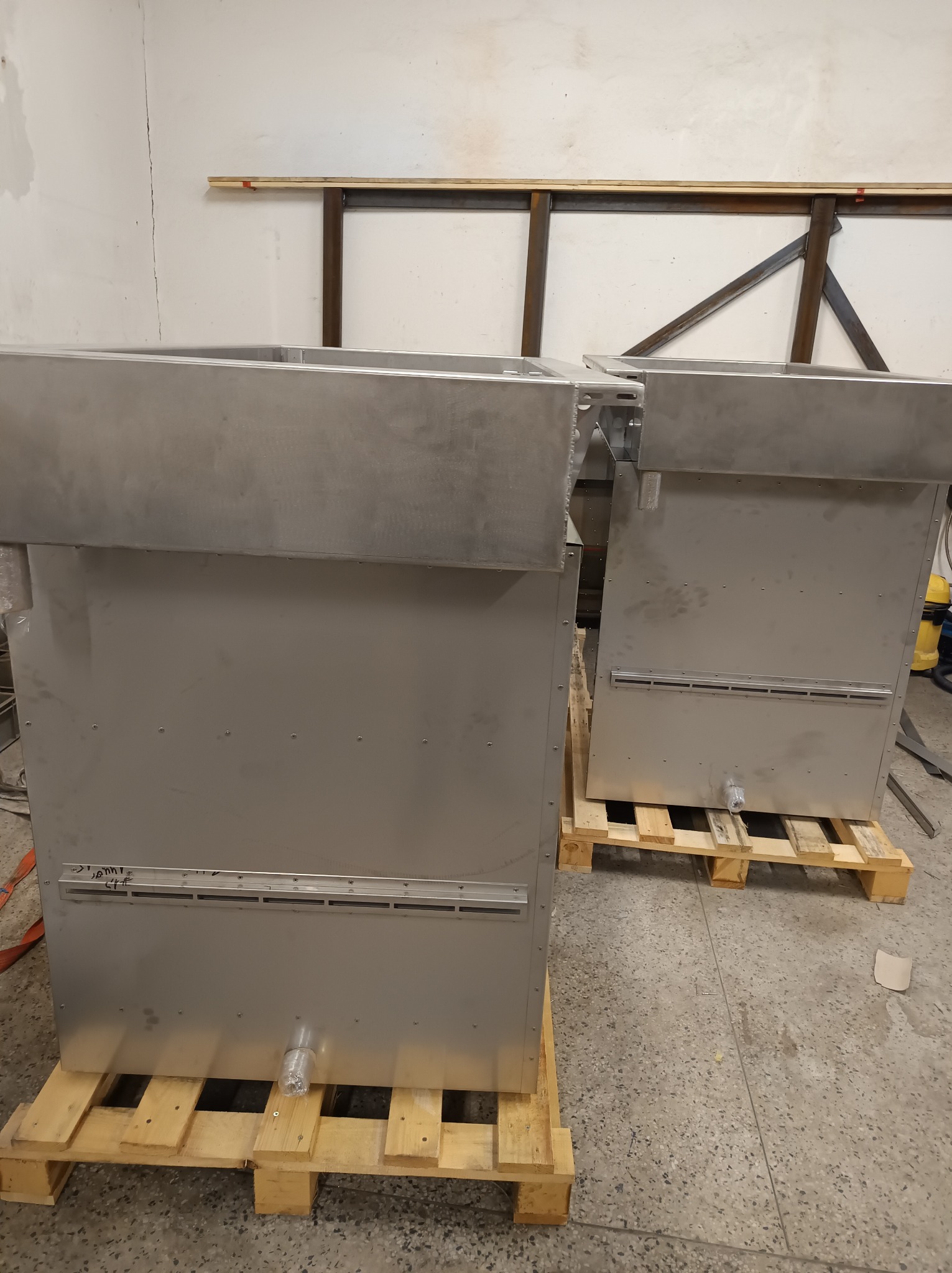 Dwie metalowe konstrukcje na paletach w warsztacie. Widoczne spawane elementy i otwory wentylacyjne. Szlifowana powierzchnia metalu.