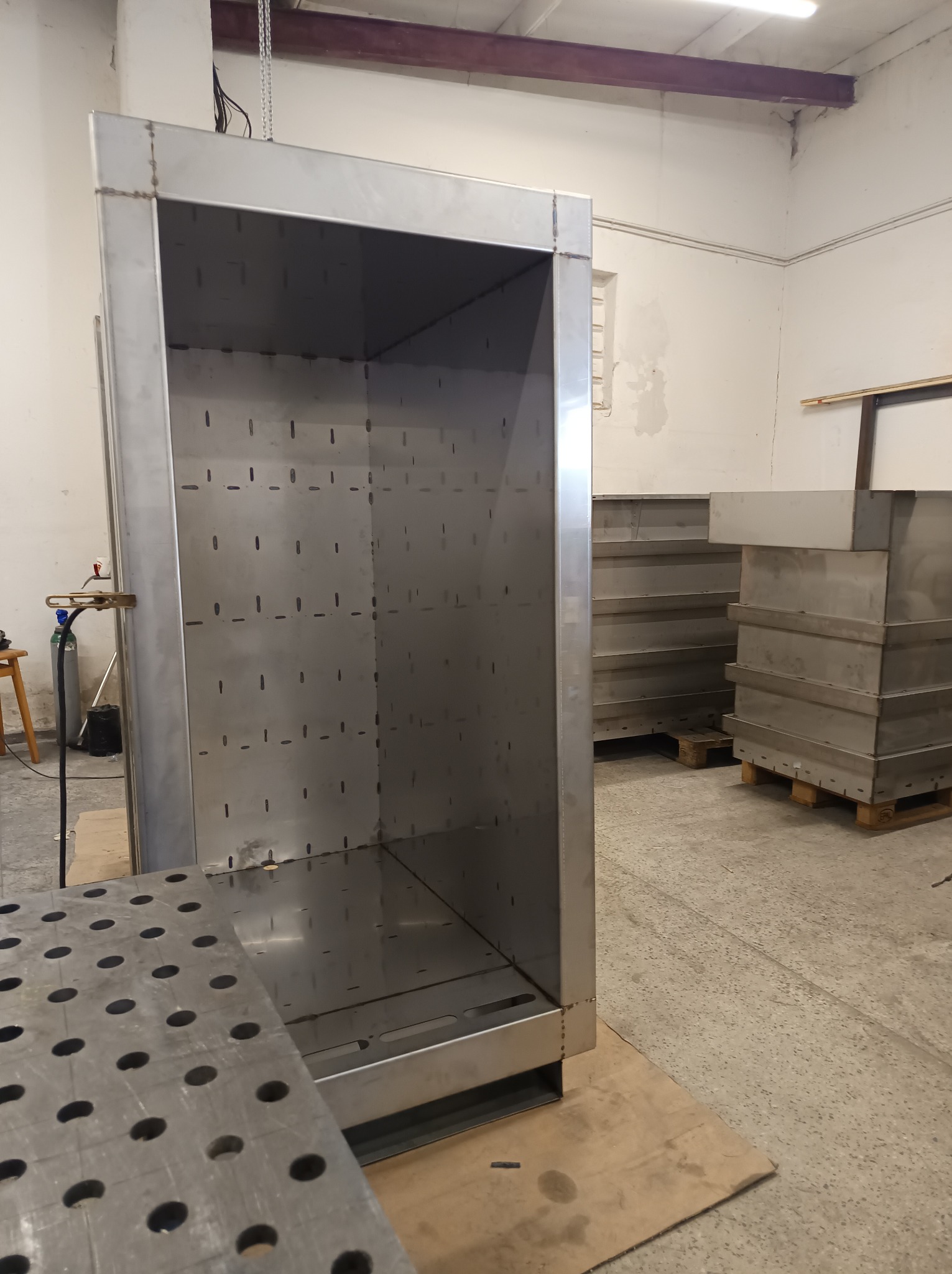 Metalowa konstrukcja szafy z otworami, spawana, w warsztacie. Widoczne narzędzia i elementy konstrukcyjne. Surowe wykończenie, przemysłowy charakter.