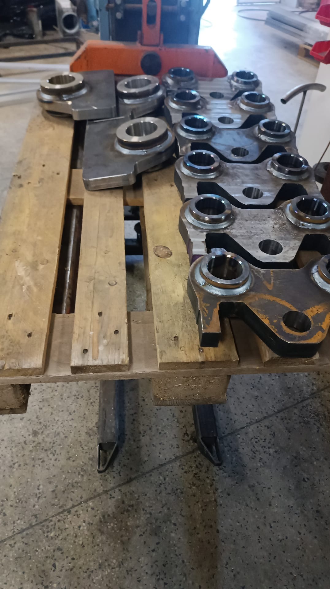 Metalowe elementy po obróbce CNC, ułożone na drewnianej palecie. Widoczne ślady obróbki i różnice w wykończeniu powierzchni. Elementy o różnych kształtach i rozmiarach.