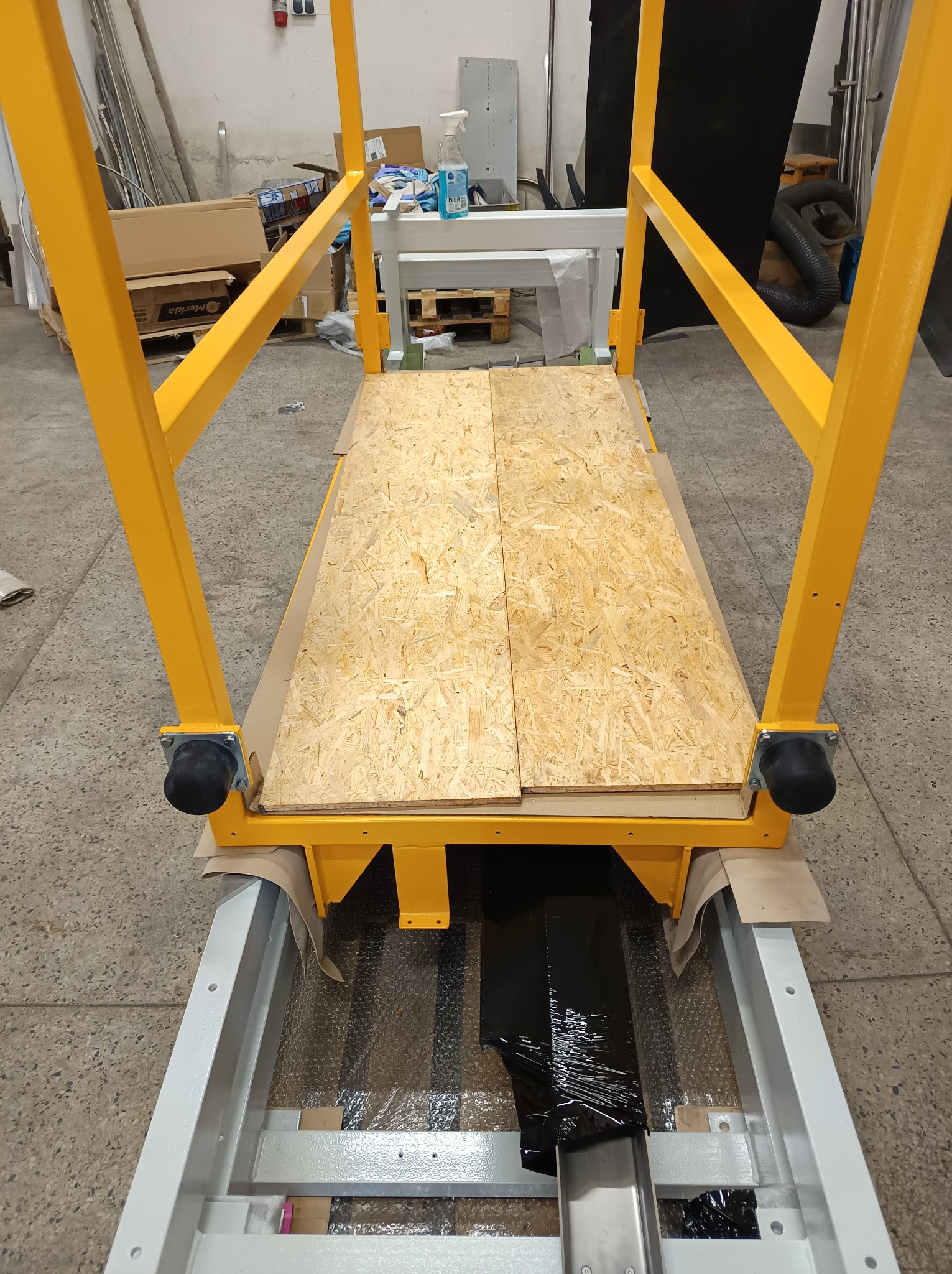 Żółta metalowa konstrukcja z drewnianym blatem na kółkach, widok z góry, w tle elementy warsztatu. Konstrukcja w trakcie montażu lub transportu.