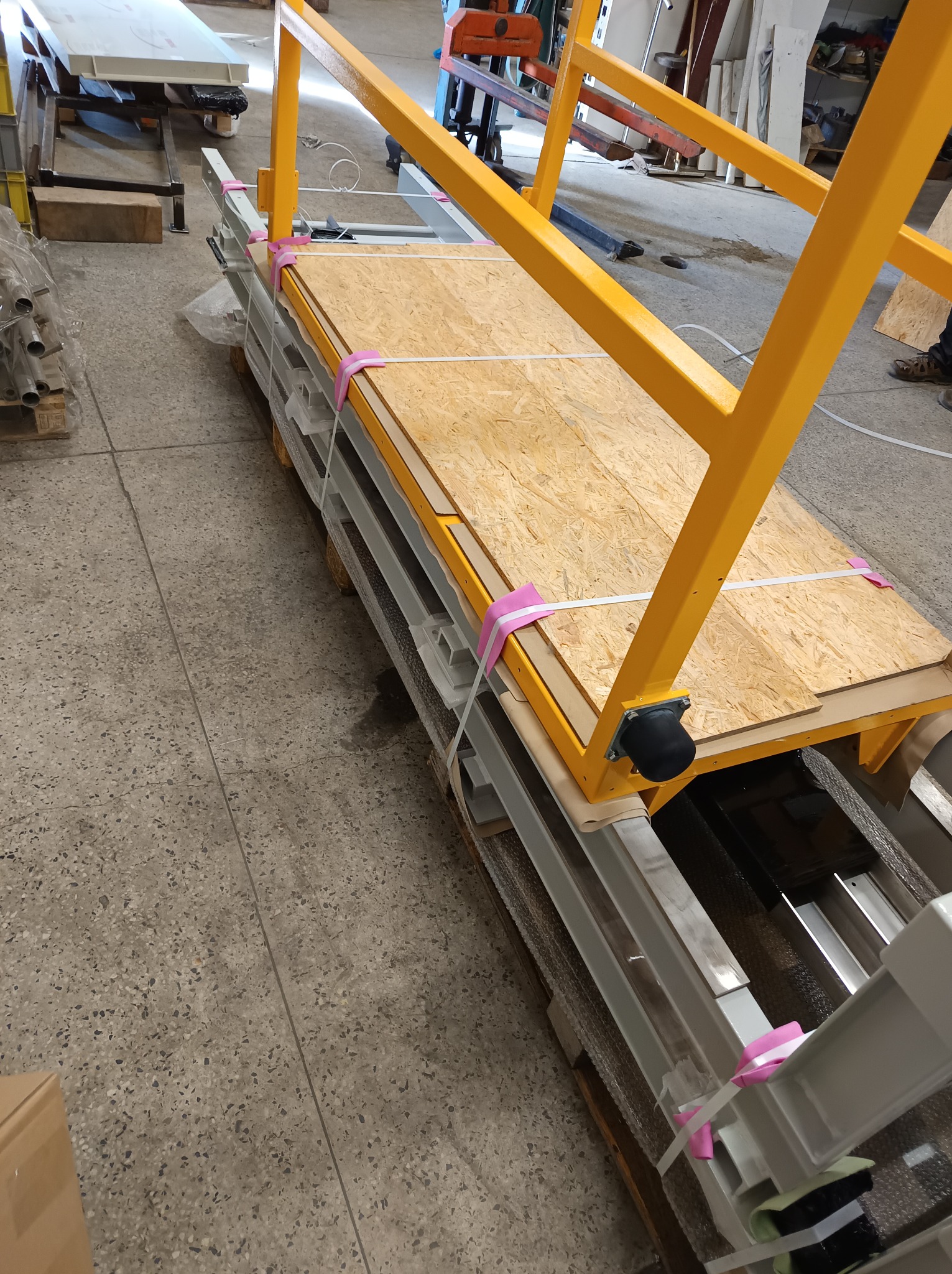 Elementy konstrukcyjne z metalu i drewna, zabezpieczone różowymi taśmami, prawdopodobnie przygotowane do transportu lub montażu. Widoczne detale spawów i łączeń.