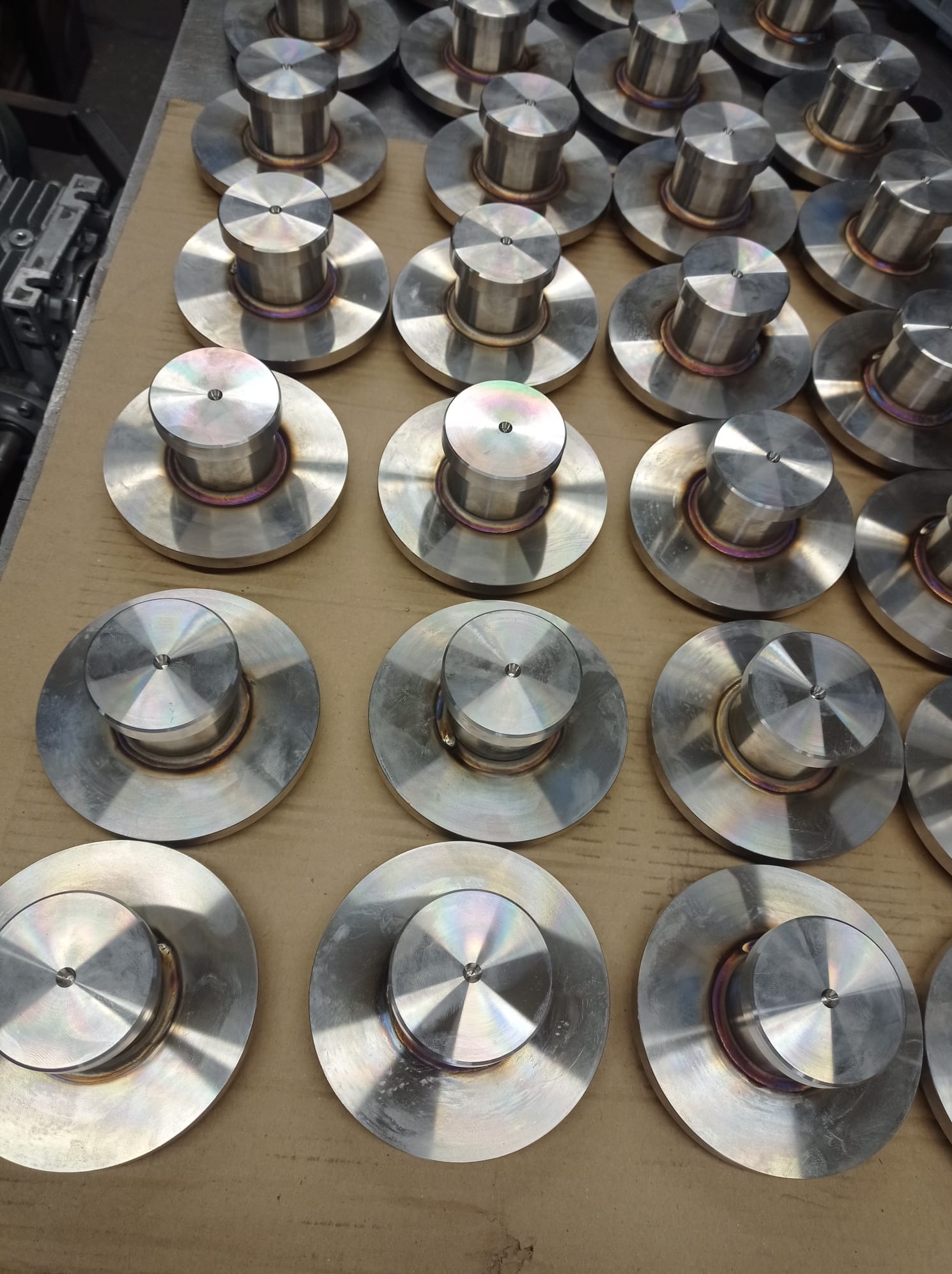 Rząd metalowych, spawanych elementów z okrągłą podstawą i cylindrycznym trzpieniem, ułożonych na kartonowym podłożu. Widoczne ślady spawania i obróbki.