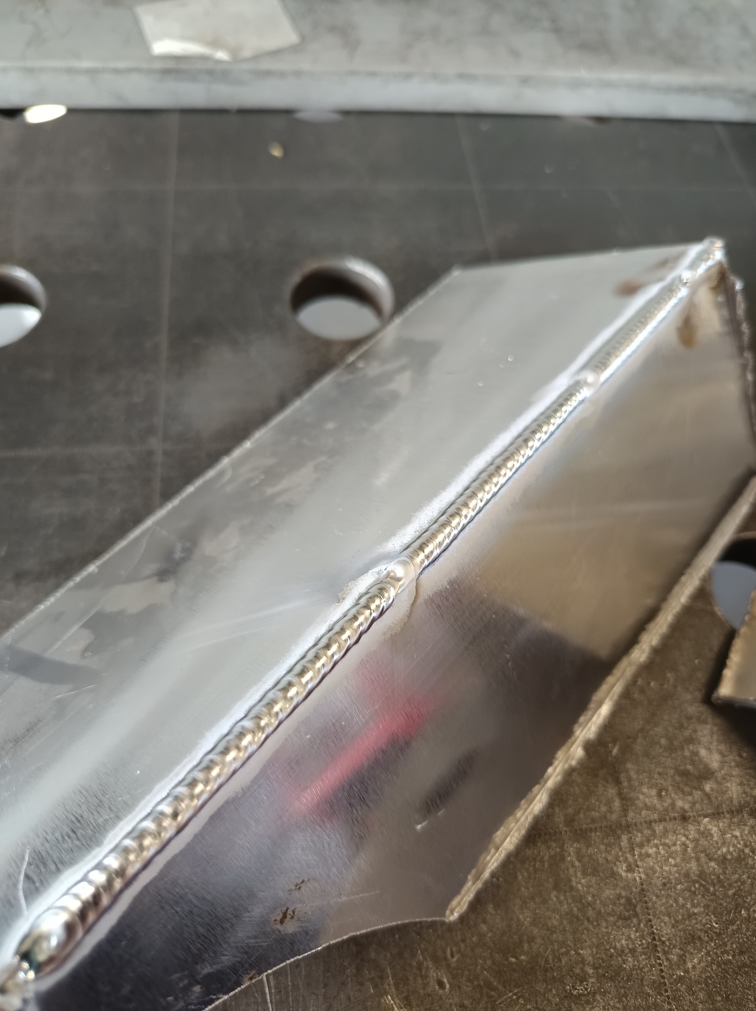 Metalowy element po spawaniu, widoczny precyzyjny spaw na krawędziach. Powierzchnia metalu odbija światło, ukazując detale obróbki. Tło stanowią narzędzia warsztatowe.