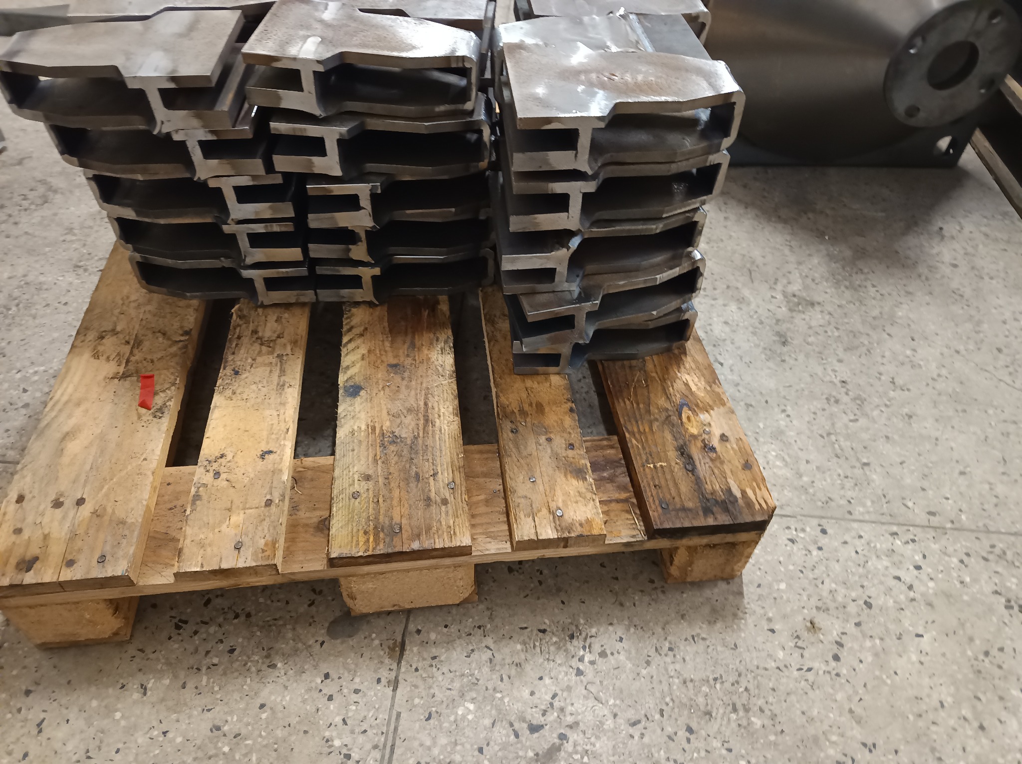 Stos metalowych profili o przekroju T, ułożonych na drewnianej palecie. Widoczne ślady obróbki. Szlifowana powierzchnia. Elementy konstrukcyjne w surowym stanie.