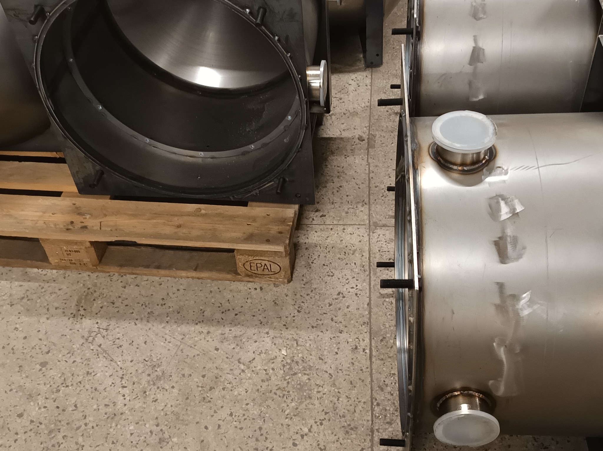 Części metalowe: czarna, okrągła rama na palecie i stalowe zbiorniki z zaślepkami, gotowe do dalszej obróbki lub montażu. Widoczne spawy i śruby.