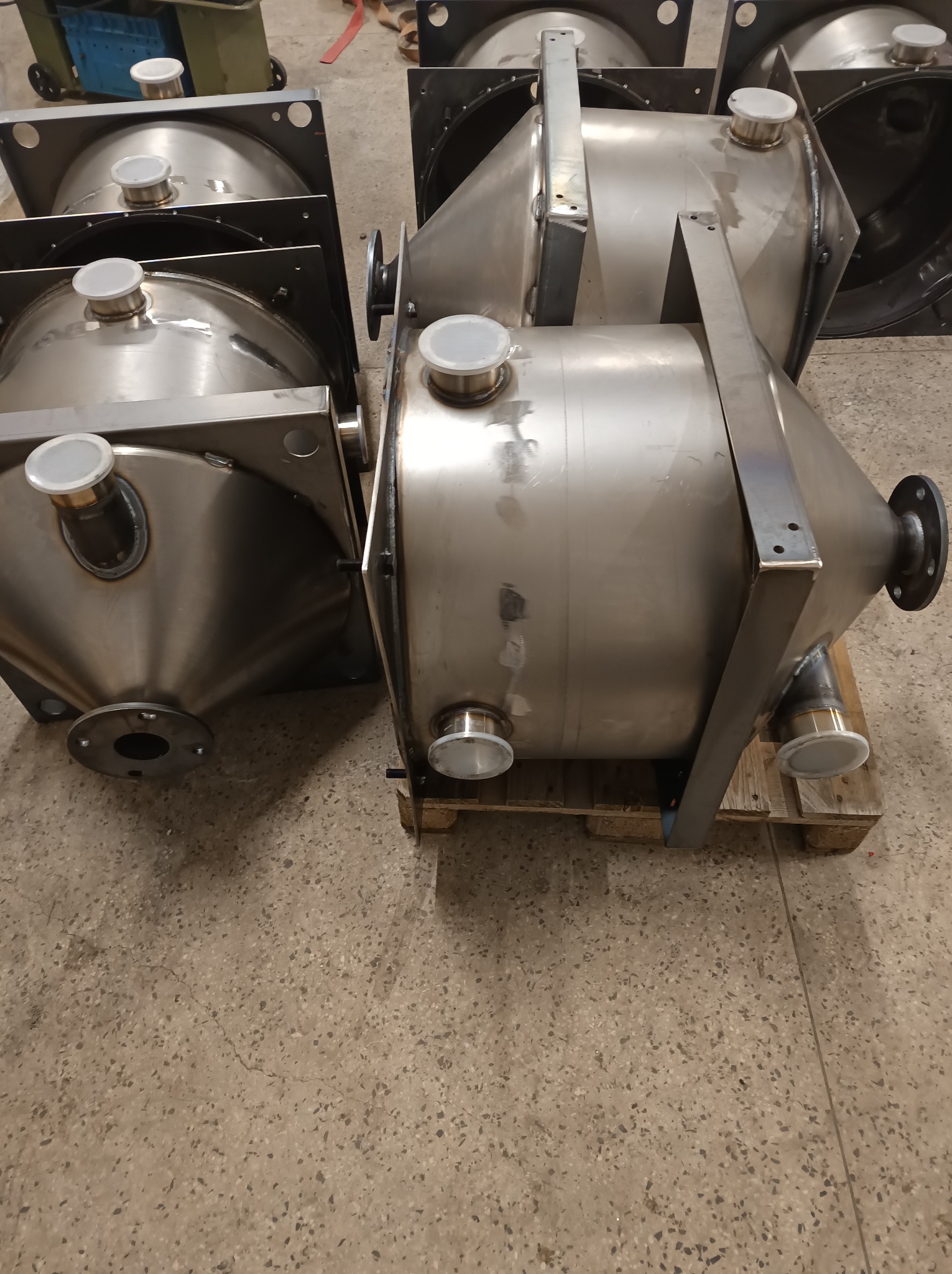 Metalowe zbiorniki o stożkowym kształcie, ustawione na palecie w hali produkcyjnej. Widoczne spawy i elementy mocujące. Wykończenie ze stali nierdzewnej.