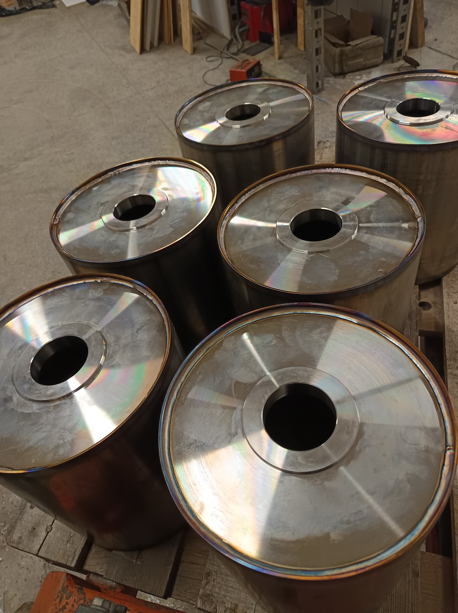 Sześć metalowych, cylindrycznych elementów z otworem centralnym, ułożonych na drewnianej palecie. Widoczne ślady spawania i opalizujące kolory na krawędziach.