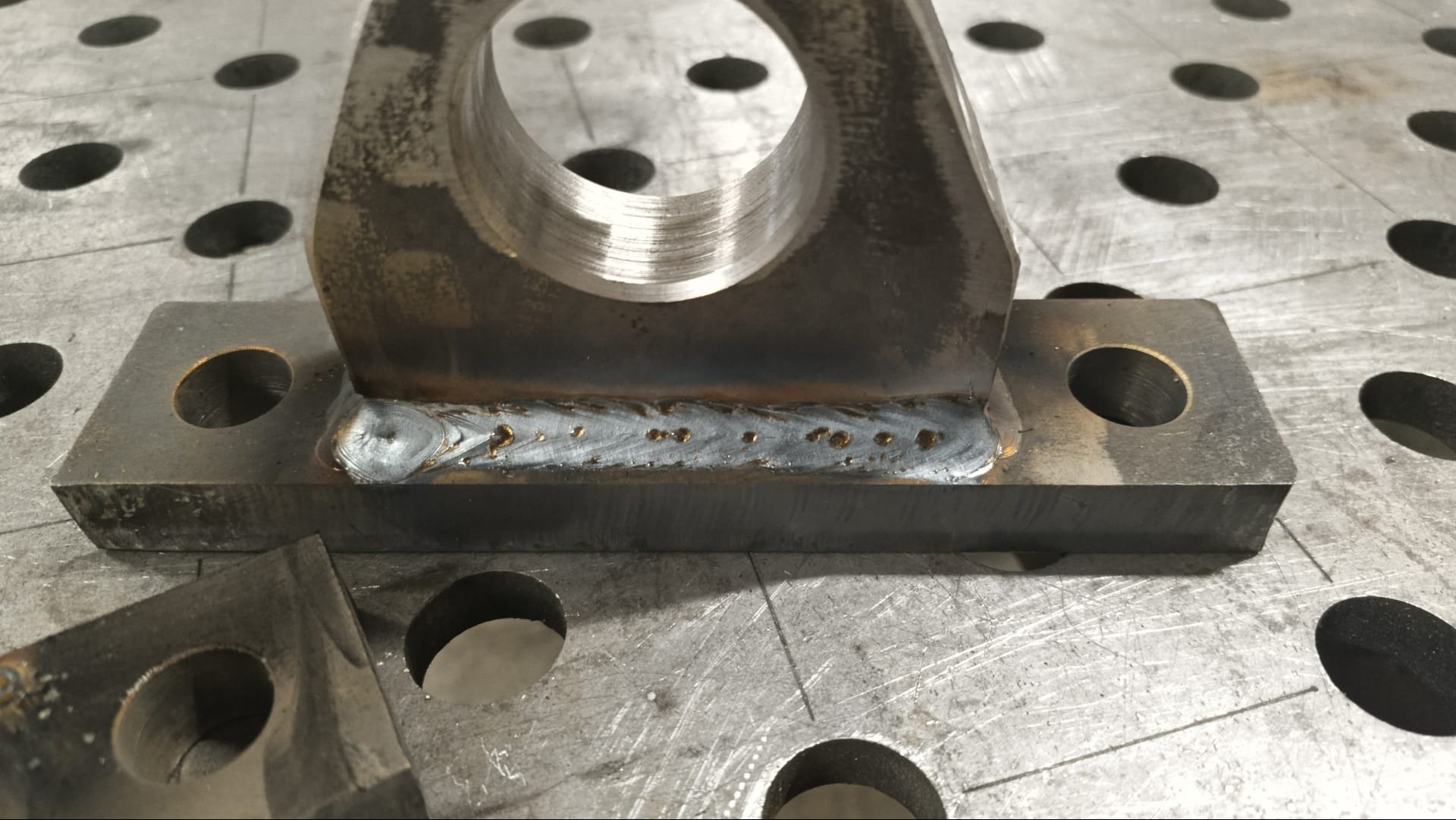 Metalowy element z widocznym spoiwem spawalniczym na perforowanym stole spawalniczym. Widoczne ślady obróbki i niedoskonałości spoiny.