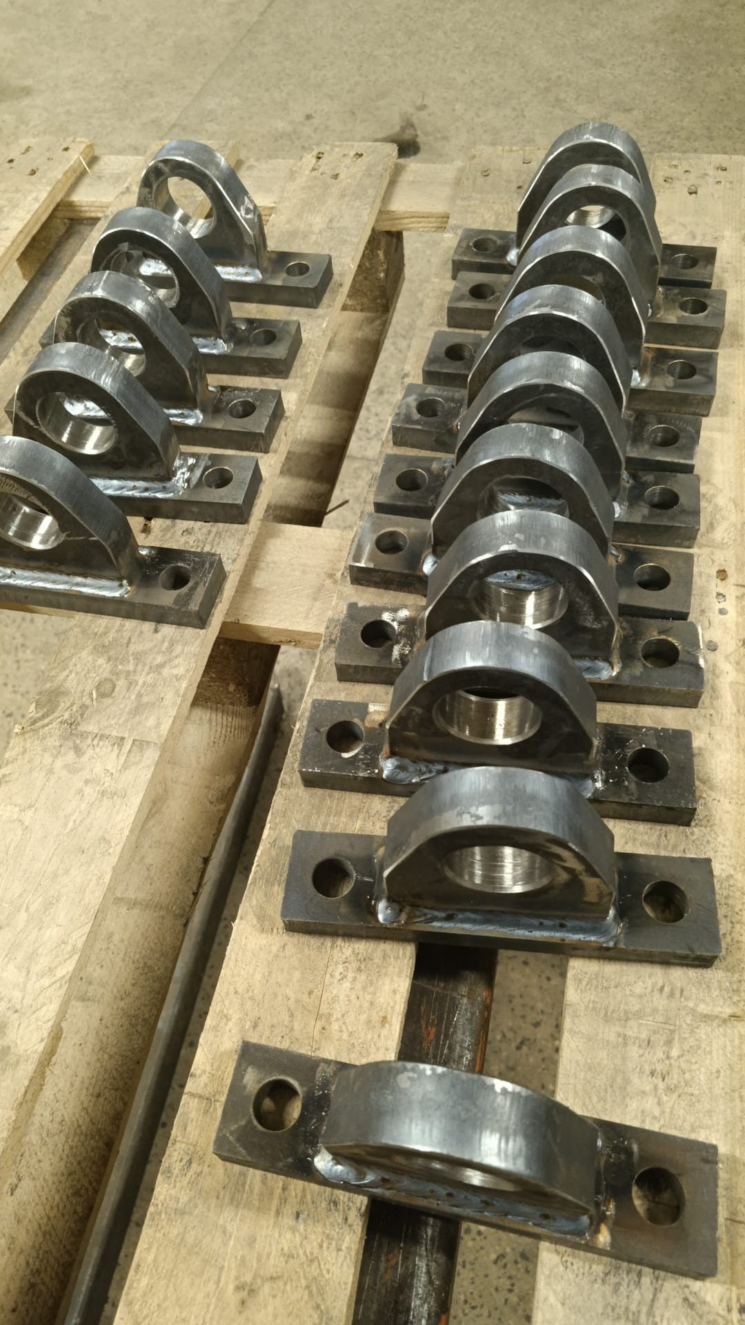 Rząd metalowych uchwytów z otworami, spawanych do płaskowników, ułożonych na drewnianej palecie. Widoczne spawy i surowy metal. Detal obróbki metalu.