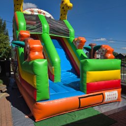 Dmuchane szaleństwo - Kolorowy dmuchany zamek ze zjeżdżalnią, motyw zwierzęcy (żyrafy, małpki), na trawie i kostce brukowej, błękitne niebo w tle. Idealny na imprezy plenerowe dla dzieci.
