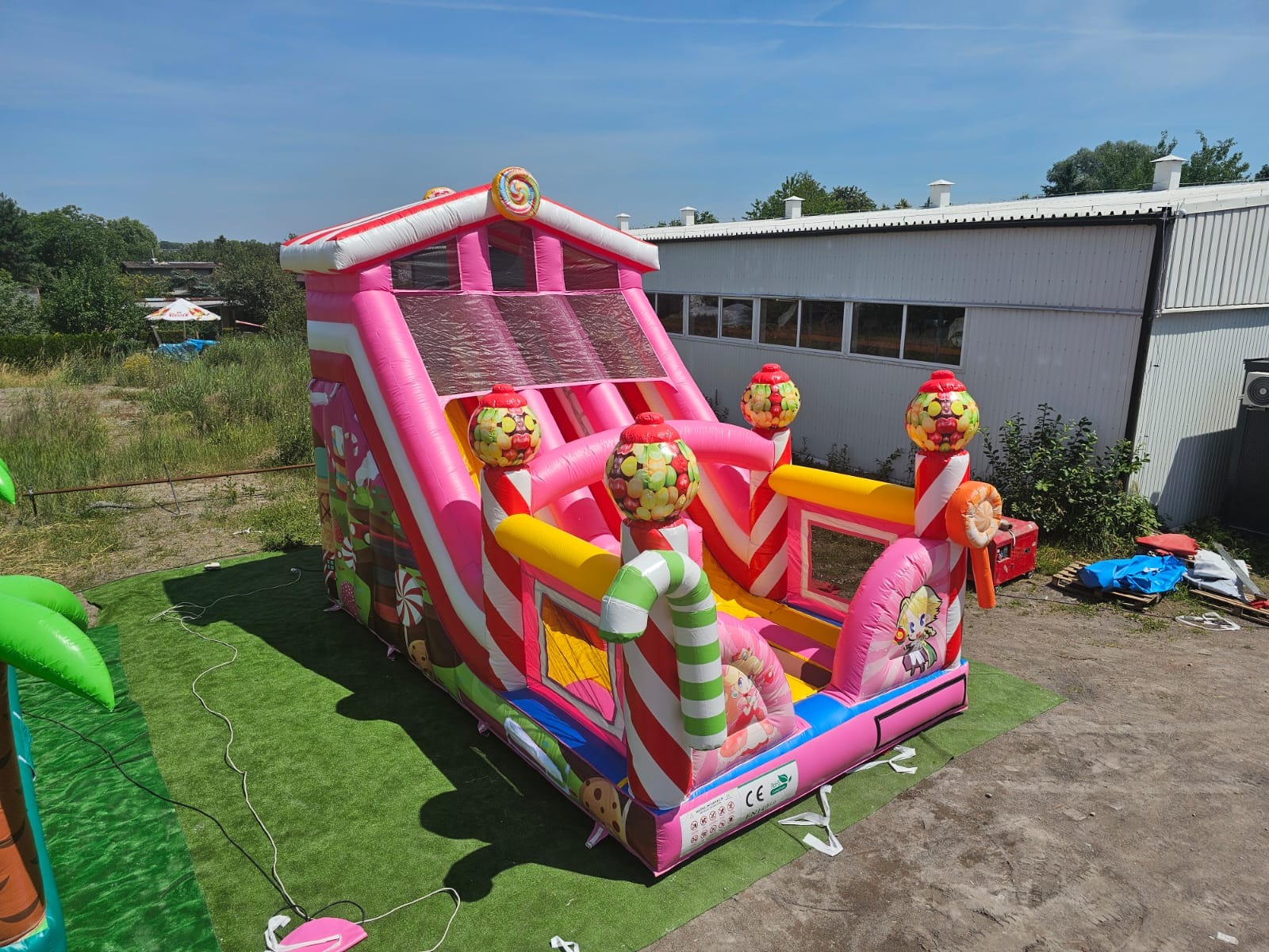 Kolorowa, dmuchana zjeżdżalnia w stylu cukierkowym na sztucznej trawie. Zjeżdżalnia ma motywy lizaków i cukierków, a w tle widoczny budynek.