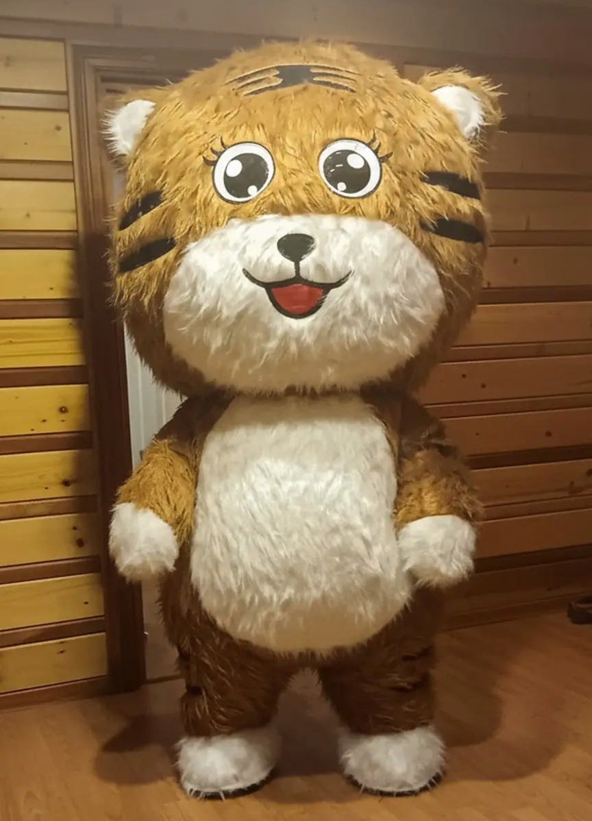 Pluszowy kostium tygryska z dużymi oczami i uśmiechem, idealny dla animatora dziecięcego na imprezę. Brązowo-biały strój na tle drewnianej ściany i drzwi.