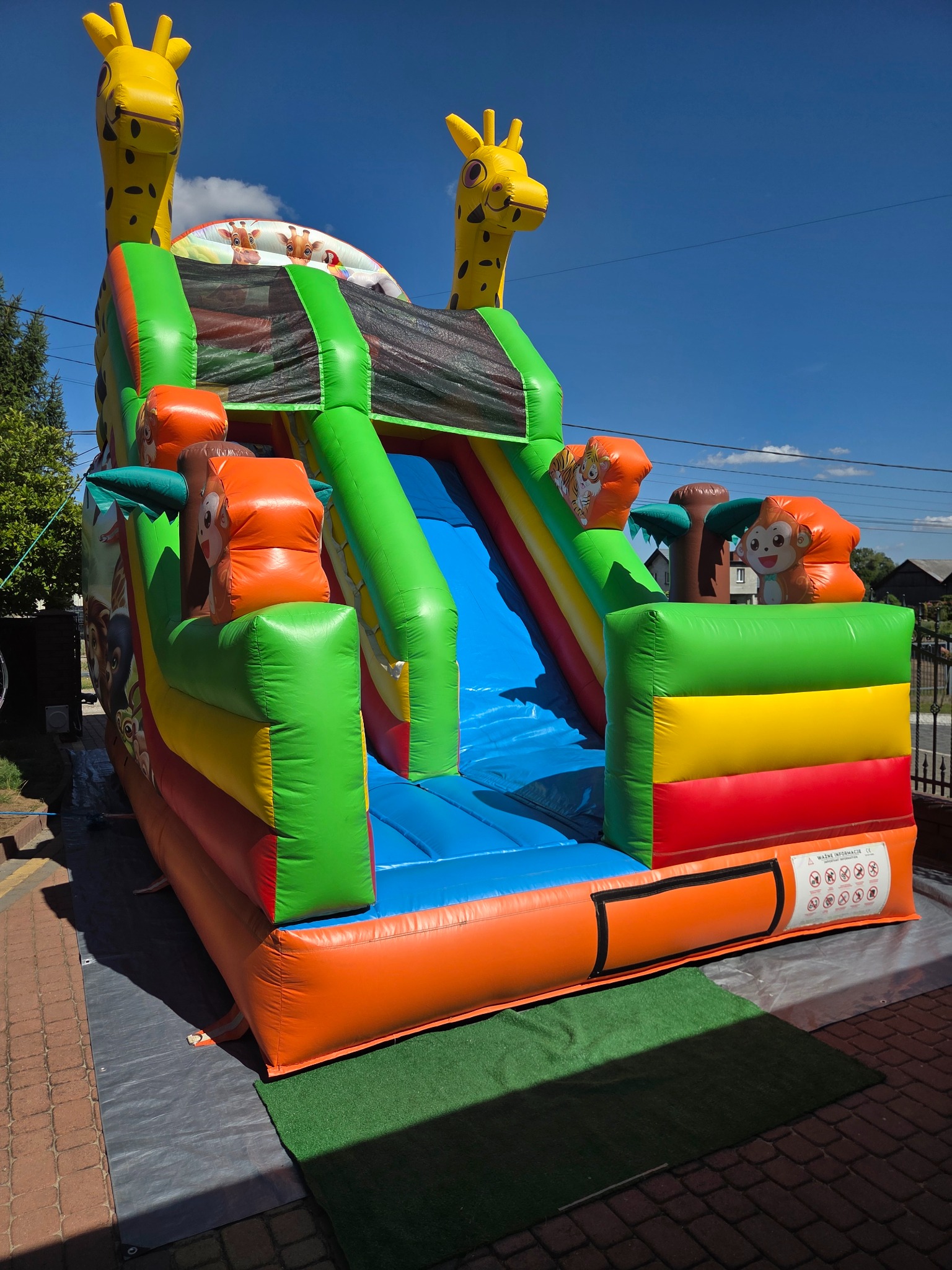 Kolorowy dmuchany zamek ze zjeżdżalnią, motyw zwierzęcy (żyrafy, małpki), na trawie i kostce brukowej, błękitne niebo w tle. Idealny na imprezy plenerowe dla dzieci.