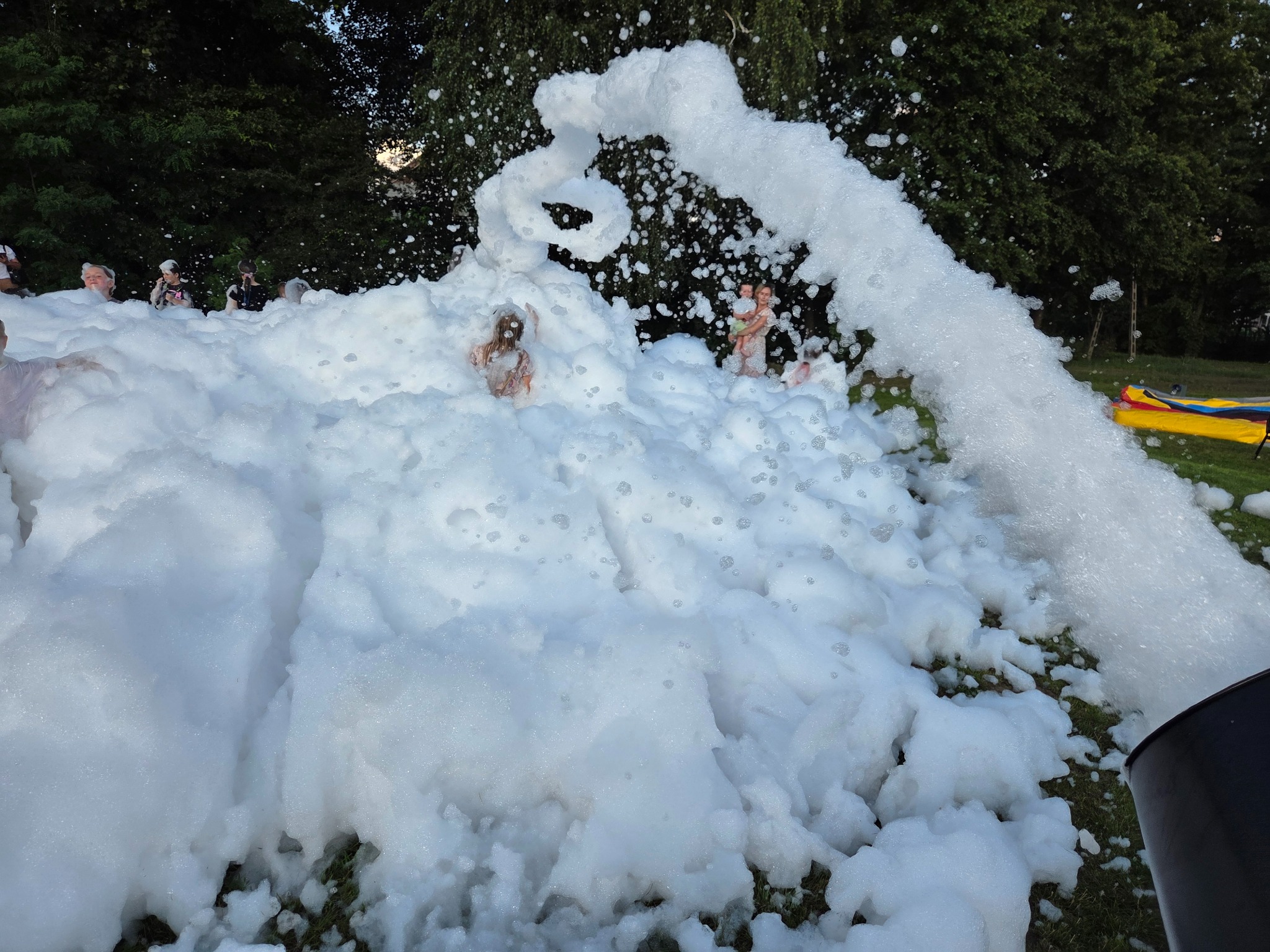 Piana party w plenerze: dzieci i dorośli bawią się w białej pianie, z dyszy wylatuje strumień piany, w tle zieleń drzew. Atrakcja na imprezę.