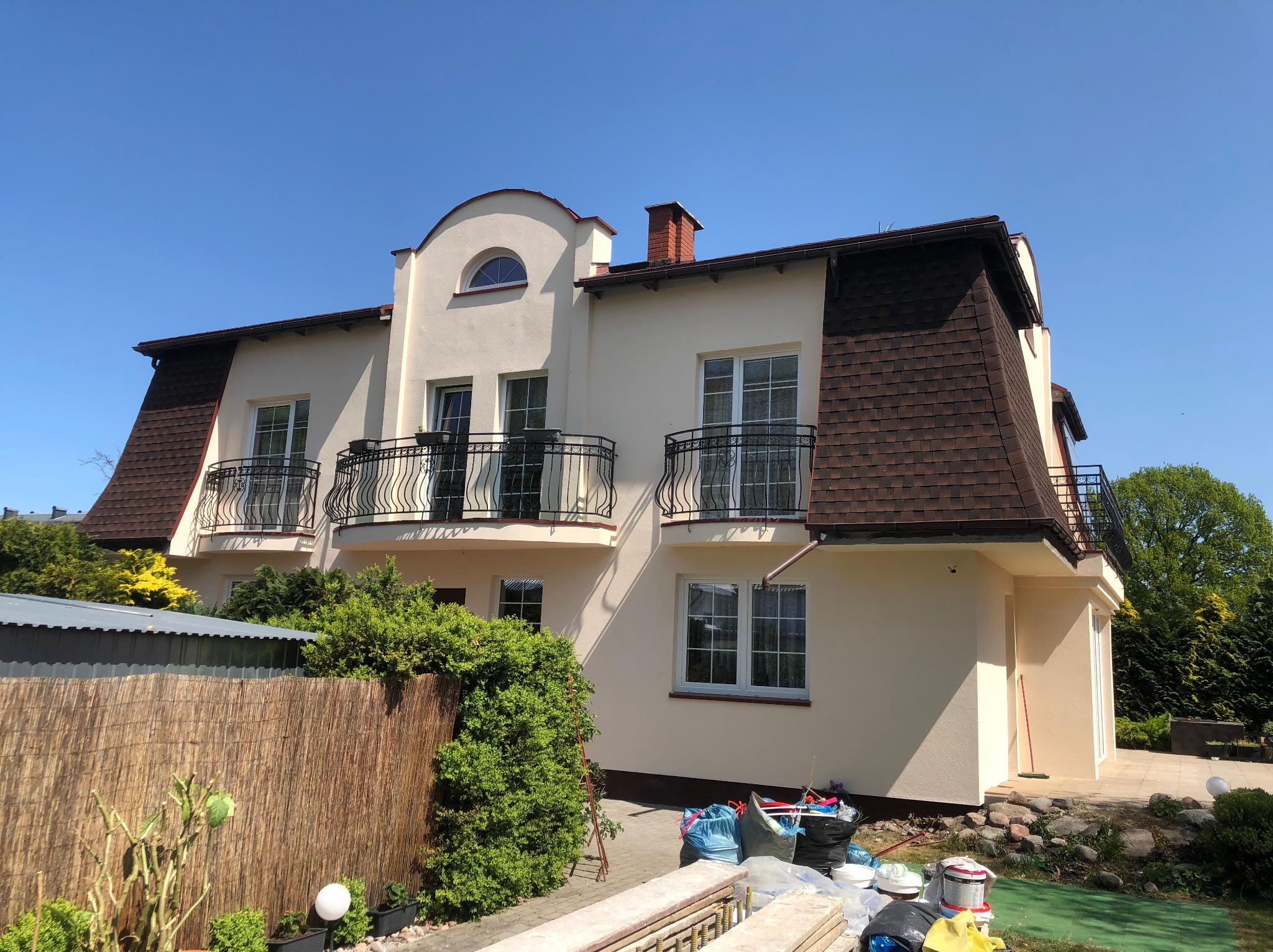 Jasny, dwupiętrowy dom z balkonami i brązowym dachem po renowacji elewacji. Widoczne detale architektoniczne i świeżo pomalowana fasada w słoneczny dzień.