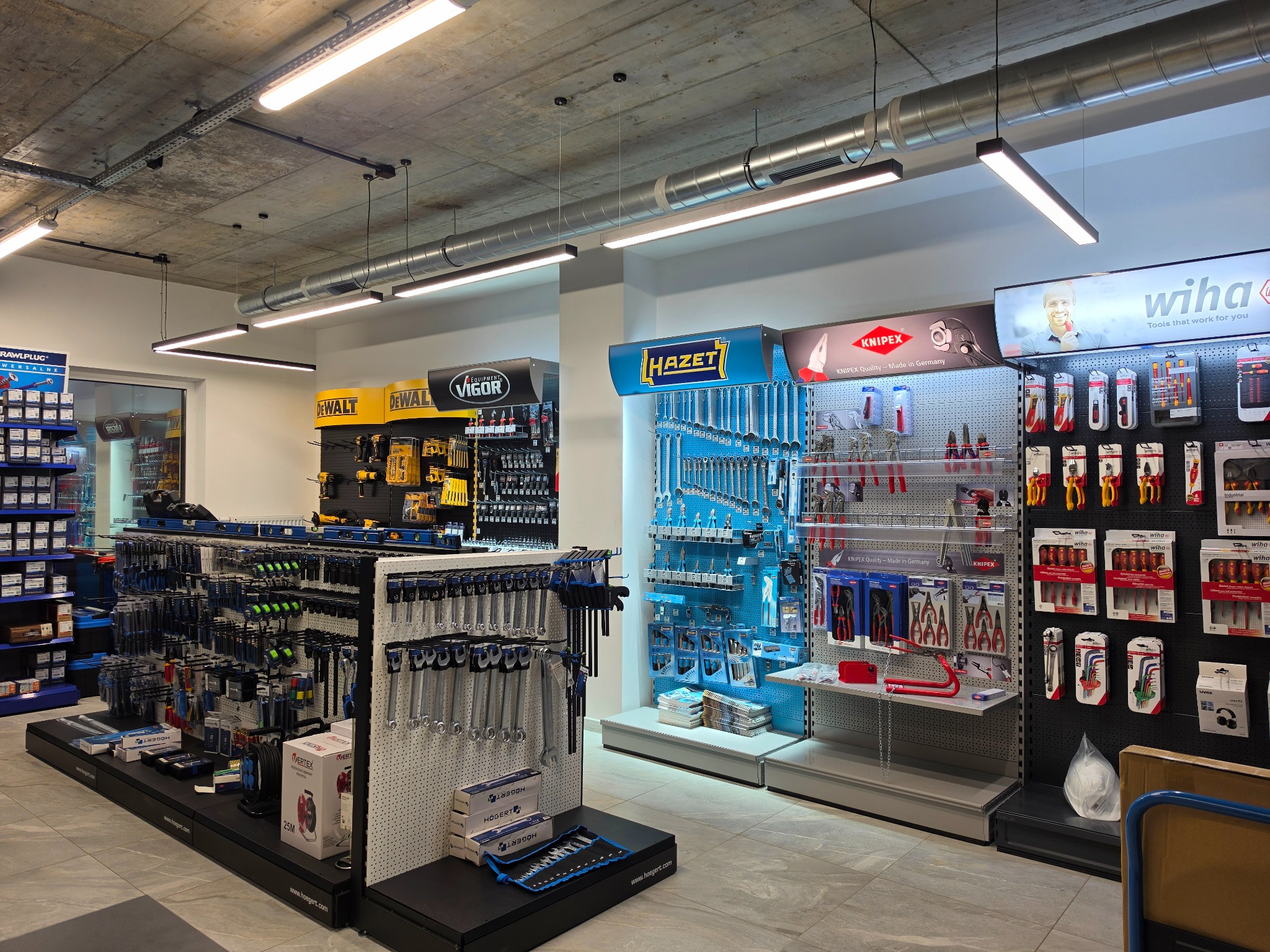 Wnętrze sklepu z narzędziami: ekspozycja marek DeWalt, Hazet, Knipex i Wiha. Różnorodność narzędzi na półkach, oświetlenie sufitowe, betonowy sufit.