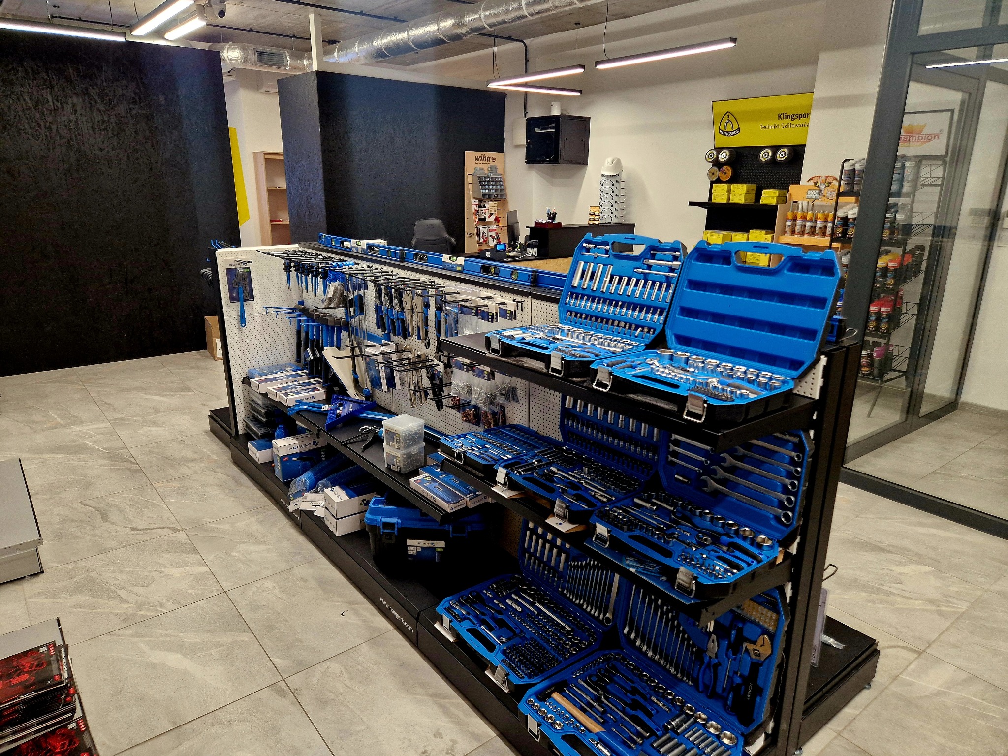 Ekspozycja narzędzi Hogert w sklepie: zestawy kluczy, wkrętaków i akcesoriów w niebieskich walizkach na czarnych regałach. Widoczne logo Wiha i Klingspor.