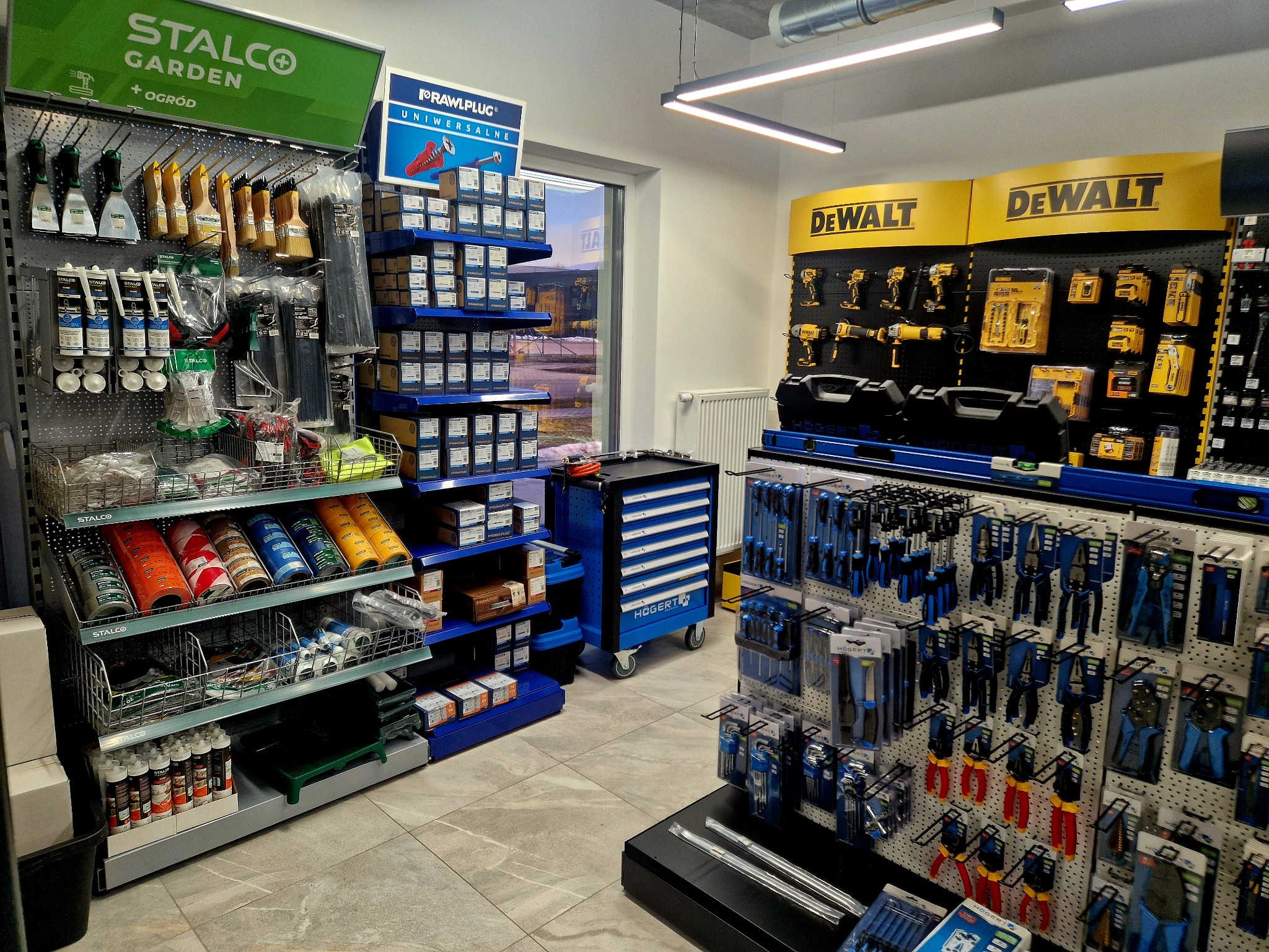 Ekspozycja narzędzi i akcesoriów budowlanych w sklepie: Stalco, Rawlplug, DeWalt, Hogert. Różnorodność produktów na półkach i wieszakach.