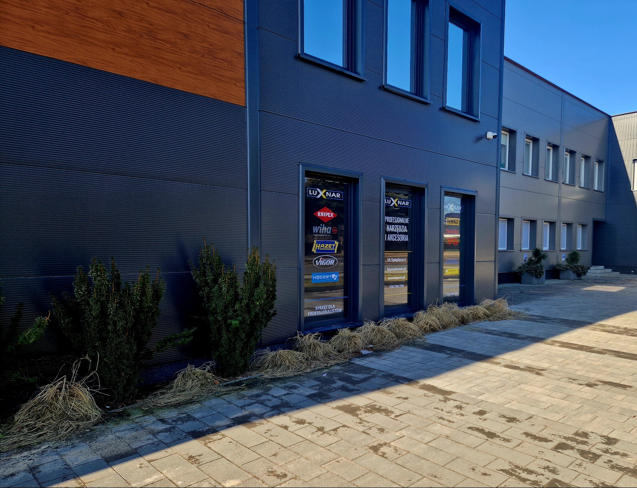 Nowoczesny budynek firmy Luxnar z logo marek narzędzi (Knipex, Wiha, Hazet, Vigor, Hogert) na witrynach. Widok z zewnątrz, kostka brukowa, krzewy.