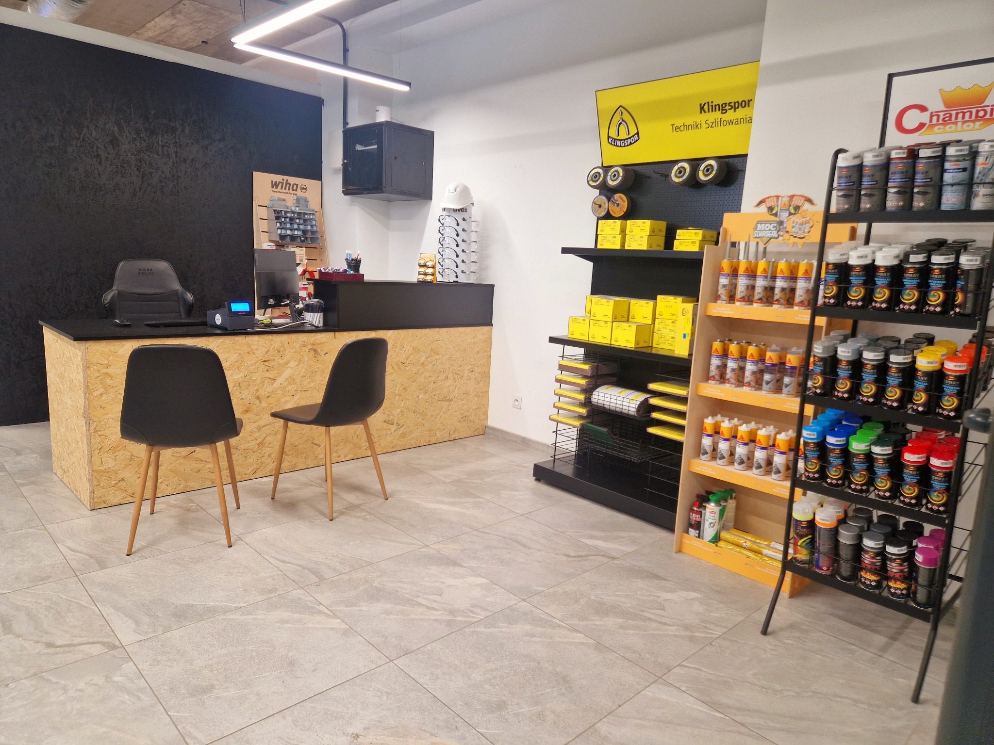 Wnętrze sklepu z narzędziami i materiałami budowlanymi: regały z farbami, szlifierkami, stanowisko obsługi klienta z krzesłami. Widoczne logo Wiha i Klingspor.
