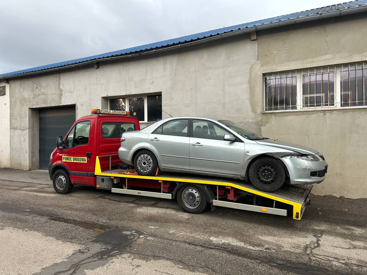 Srebrny samochód z uszkodzonym przodem na żółtej lawecie pomocy drogowej, na tle szarego budynku z oknami w kraty i czerwonego pojazdu z napisem 'Pomoc Drogowa'.