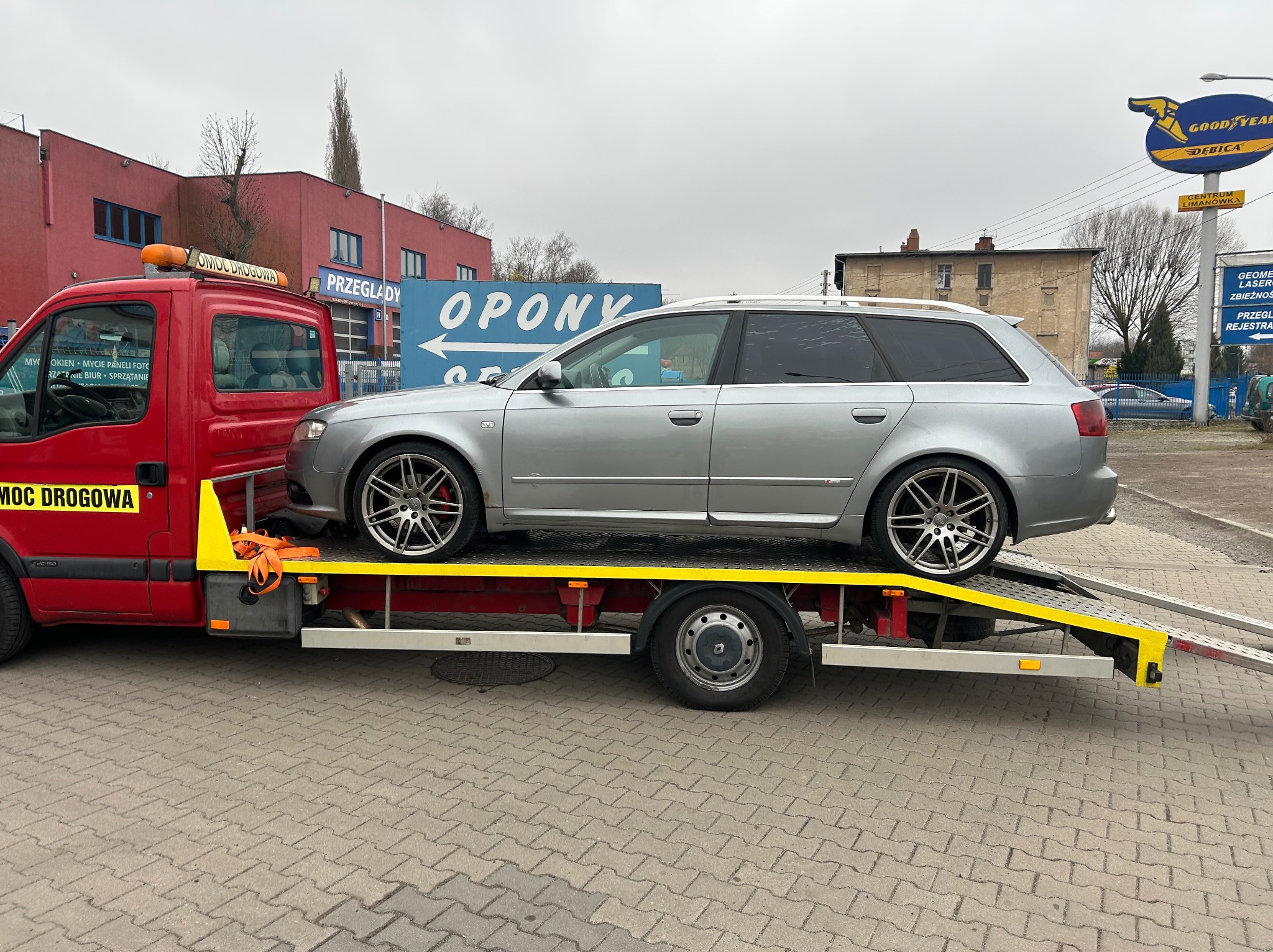 Srebrne kombi Audi na żółtej platformie czerwonej lawety pomocy drogowej, na tle budynków i szyldów reklamowych. Widok z boku, światło dzienne.