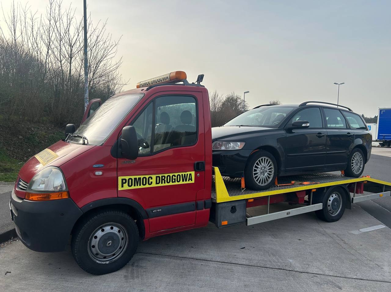 Czerwony pojazd pomocy drogowej z czarnym kombi na platformie, widoczne oznaczenia 'Pomoc Drogowa' i pomarańczowe światło ostrzegawcze na dachu.