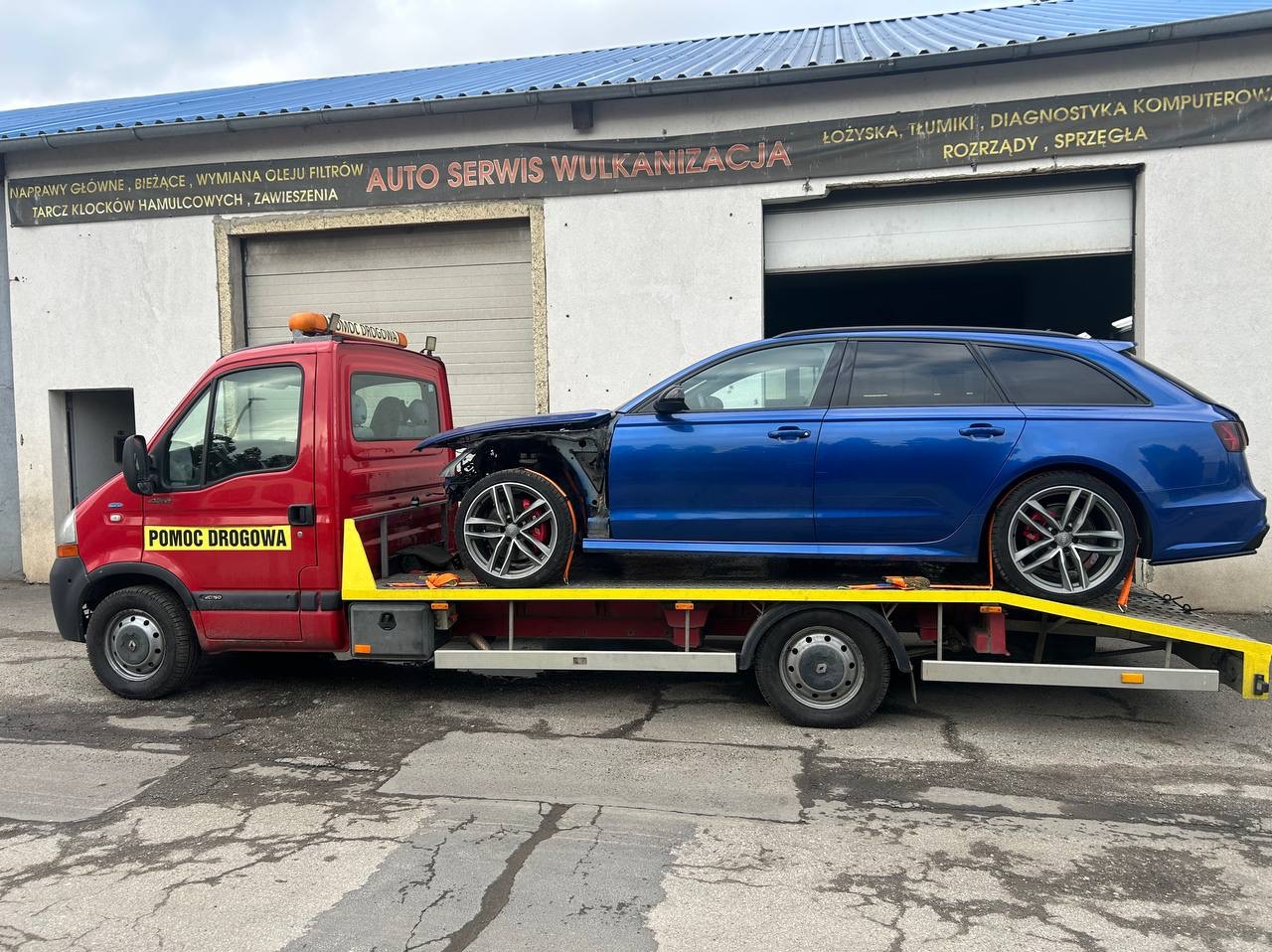 Czerwona laweta z uszkodzonym, niebieskim Audi kombi na platformie, na tle budynku auto serwisu. Widoczny brak przedniej części pojazdu.