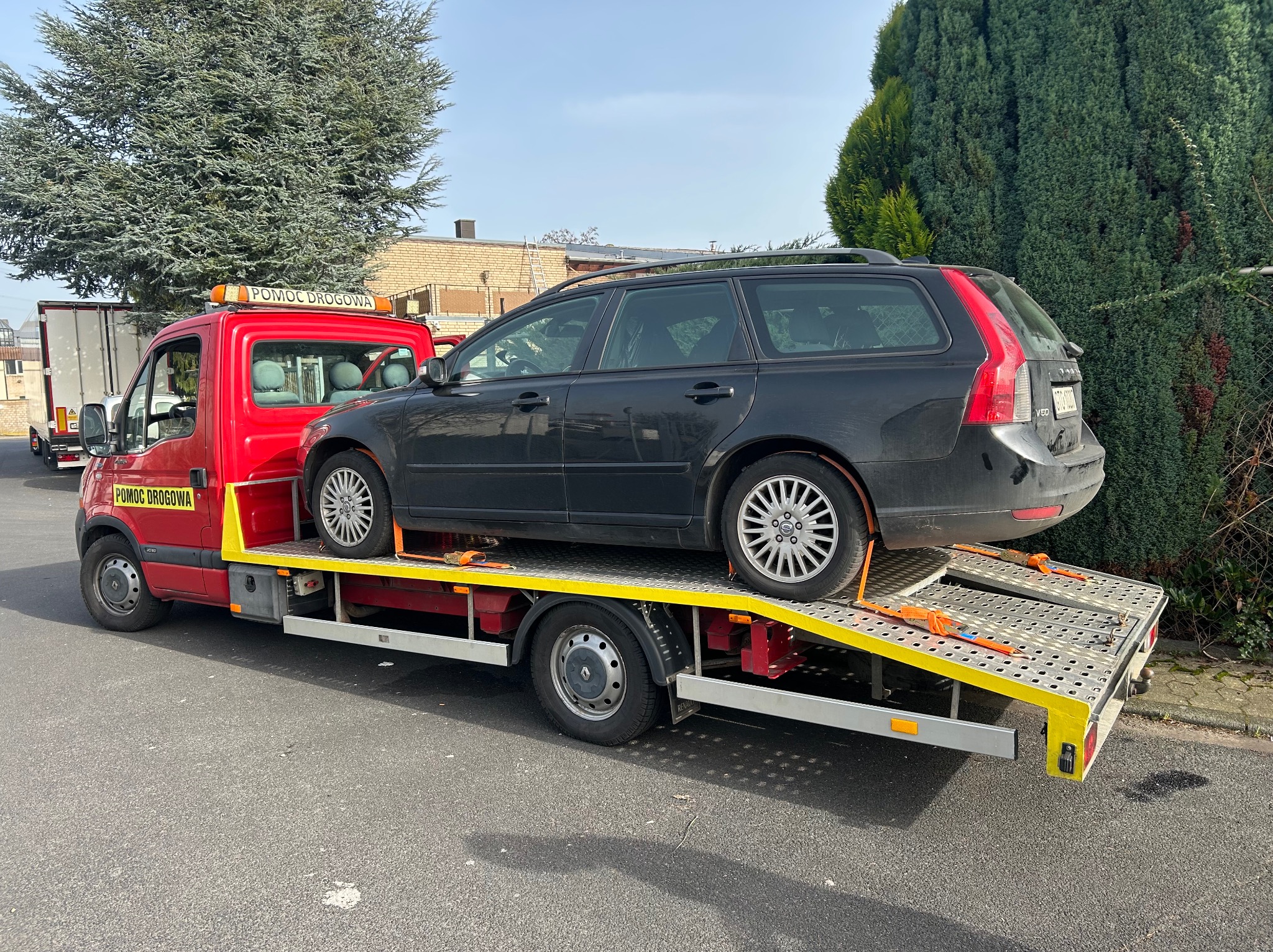 Czerwona autolaweta z napisem 'Pomoc Drogowa' przewozi czarne kombi Volvo na żółtej platformie. Widok z boku, w tle budynek i zieleń.