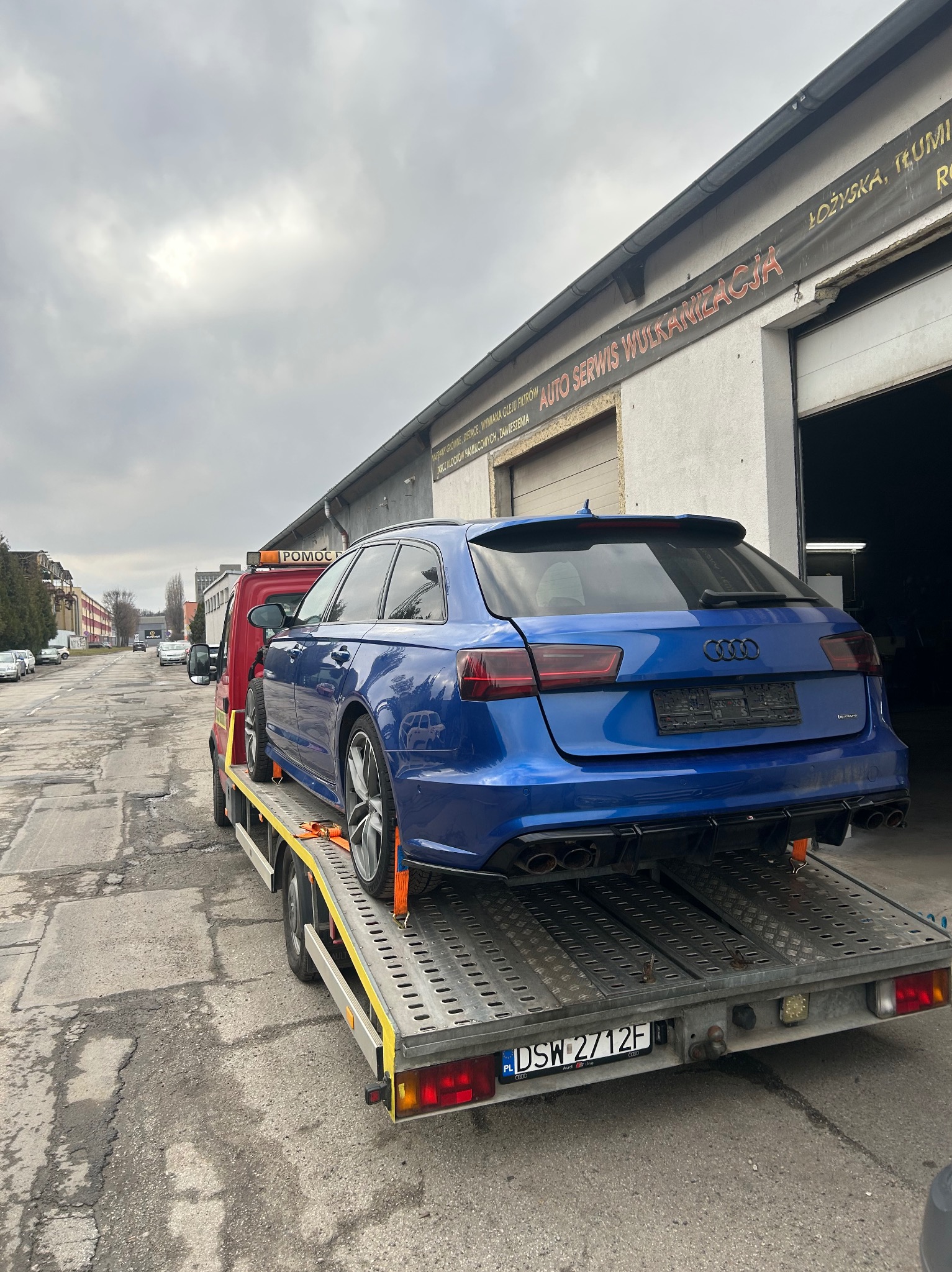 Niebieskie Audi RS6 Avant na platformie pomocy drogowej, widok z tyłu, tablica rejestracyjna DSW 2712F, w tle budynek auto serwisu z napisem 'AUTO SERWIS WULKANIZACJA'.
