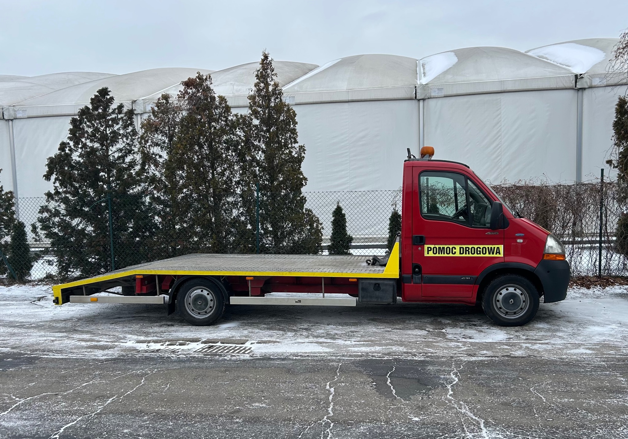Czerwona laweta z żółtymi elementami, zaparkowana na ośnieżonym placu, na tle białego namiotu i zielonych drzew. Widok z boku, pochmurny dzień.