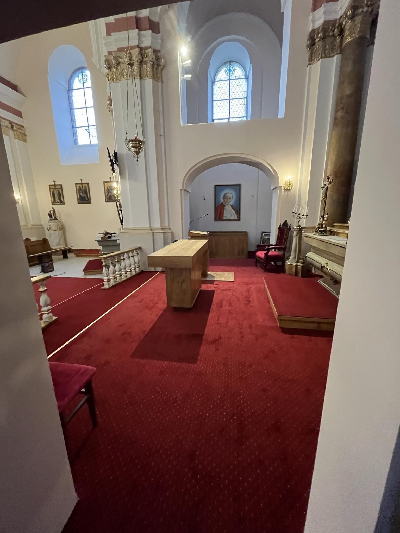 Wnętrze kościoła z czerwonym dywanem, drewnianym ołtarzem i portretem papieża w tle. Widok zza filaru, okna łukowe wpuszczają światło.