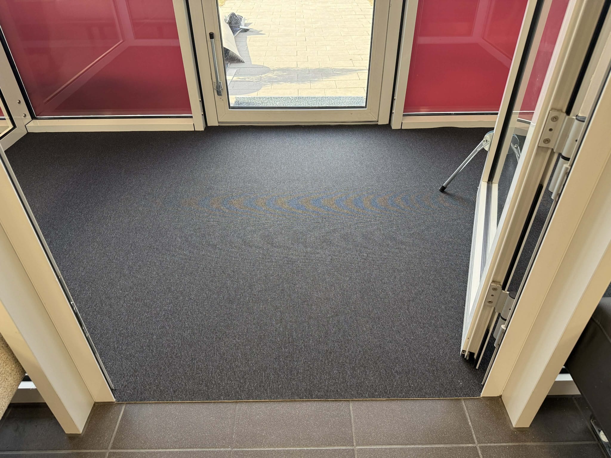 Ciemnoszara wykładzina dywanowa z subtelnym wzorem, ułożona w wejściu z białymi drzwiami i czerwonymi szybami. Widok z góry, fragment podłogi z płytek.