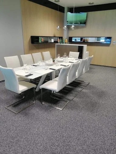 Nowoczesna sala konferencyjna z szarą wykładziną dywanową, białym stołem i krzesłami. Minimalistyczny design, oświetlenie punktowe i telewizor na ścianie.