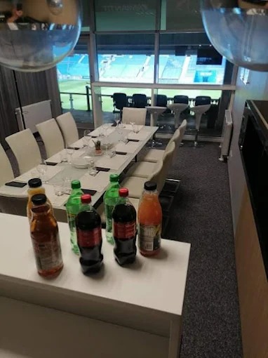 Nowoczesne wnętrze loży VIP z widokiem na stadion. Długi stół z białymi krzesłami, napoje na blacie. Szara wykładzina podłogowa. Duże okna z widokiem na trybuny.