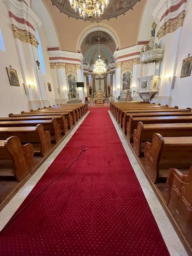 Czerwona wykładzina w kościele, biegnąca przez nawę główną. Widoczne drewniane ławki po bokach i bogato zdobione wnętrze świątyni z żyrandolami.