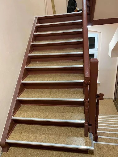 Schody z brązowymi stopniami pokrytymi wykładziną o teksturze piasku, zabezpieczone srebrnymi listwami. Brązowa balustrada po prawej stronie. Widok z dołu.