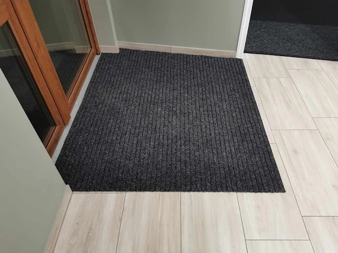 Szara, prążkowana wycieraczka podłogowa przy drewnianych drzwiach na jasnych płytkach. W tle widoczna kolejna wycieraczka w progu.
