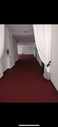 Długi korytarz z bordową wykładziną dywanową, białymi ścianami i dekoracyjnymi, białymi zasłonami po bokach. Widok w głąb korytarza.