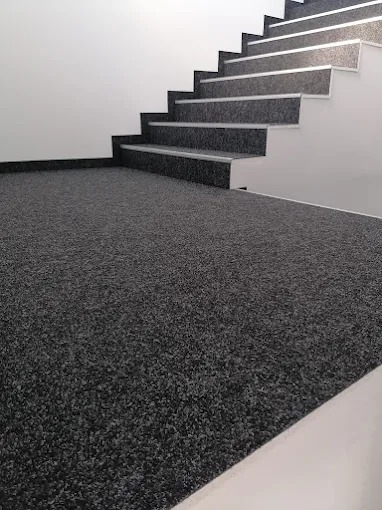 Ciemna wykładzina dywanowa na podłodze i schodach w nowoczesnym wnętrzu. Minimalistyczny design, białe ściany i listwy przypodłogowe.