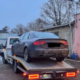 Mykyta Oleshchenko DM Company - Szary samochód osobowy marki Audi A4, z widocznymi śladami zabrudzeń, transportowany na lawecie w pochmurny dzień. Widoczny fragment zabudowań w tle.