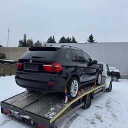 Mykyta Oleshchenko DM Company - Czarny SUV BMW na lawecie w zimowej scenerii. Auto zabezpieczone pasami transportowymi, widoczny fragment drugiego pojazdu w tle. Śnieg na ziemi i pochmurne niebo.
