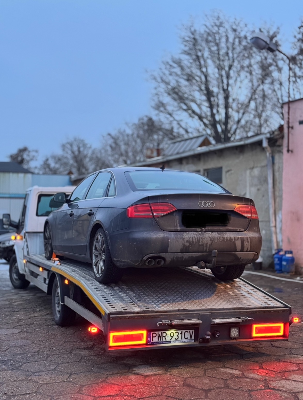 Szary samochód osobowy marki Audi A4, z widocznymi śladami zabrudzeń, transportowany na lawecie w pochmurny dzień. Widoczny fragment zabudowań w tle.